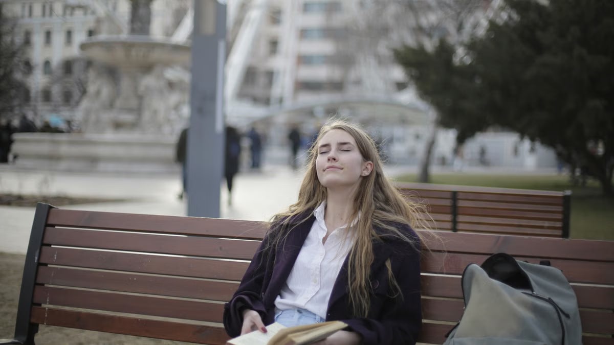 Woman enjoying a relaxed moment on a park bench with her eyes closed and a book in hand.